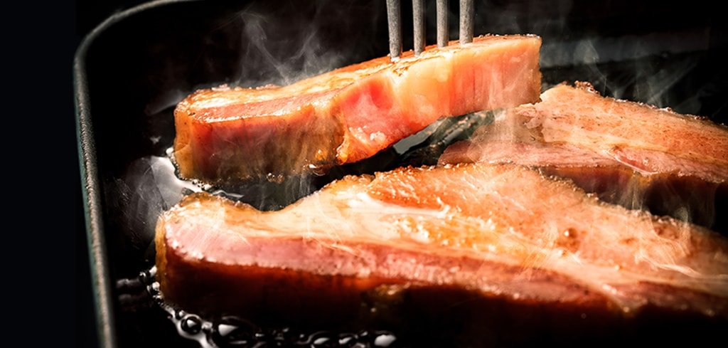 鉄板の上、フォークで持ち上げた焼きたてベーコンから湯気が立ち、脂がとろけている