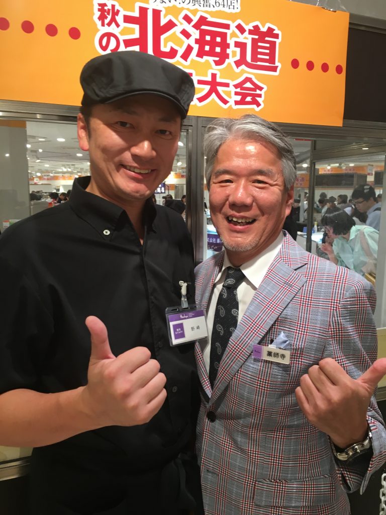 秋の北海道物産大会の看板の下で並ぶ、エーデルワイスファームの代表と阪急うめだ本店のバイヤー薬師寺氏